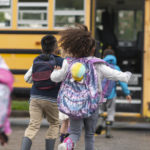 A multi-ethnic group of elementary age children are getting on a school bus. The kids' backs are to the camera. They are running towards the school bus which is parked with its door open. It's a rainy day and the kids are wearing jackets, rain boots and backpacks.